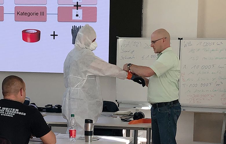 Ein Mensch in einem Schutzanzug erhält von einer Person, die Handschuhe und ein Oberteil trägt, Anweisungen zur Ausrüstung. Im Hintergrund ist eine Präsentation zu sehen, die Informationen über Sicherheitskategorien enthält. Die Szene spielt in einem Schulungsraum.